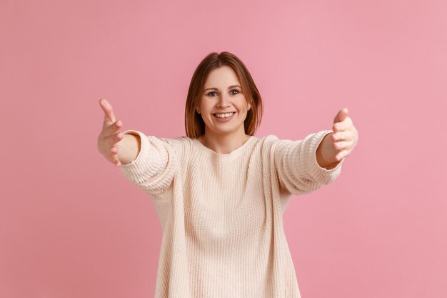 내 품으로 오세요. 하얀 스웨터를 입고 무료 포옹을 하고 환영하며 포옹을 하기 위해 손을 뻗은 웃는 금발 여성의 초상화. 분홍색 배경에 격리된 실내 스튜디오 촬영.