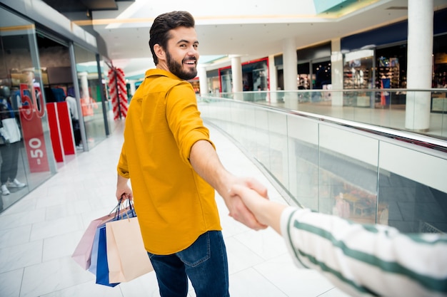 Come with me. Photo of cheerful handsome guy leading girlfriend to secret surprise place carry many bags walk shopping center toothy smiling shopper wear casual outfit indoors