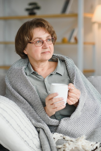 Comfort relaxed senior old elderly woman grandmother drinking\
hot beverage tea coffee at home looking at camera in the living\
room pension concept