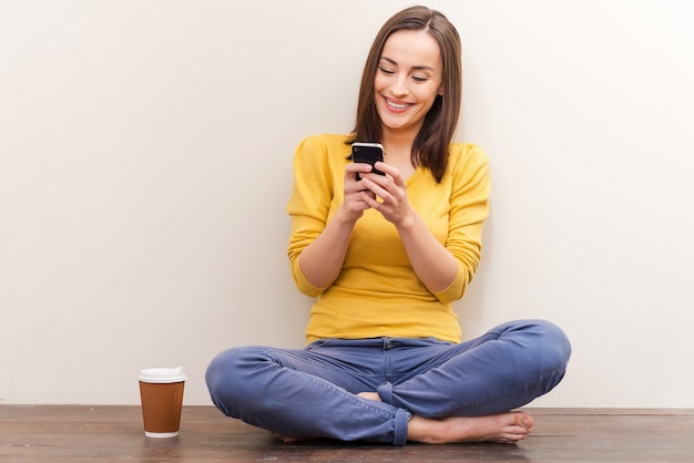 Comfortabele communicatie. Foto van een mooie jonge vrouw die een mobiele telefoon vasthoudt terwijl ze op de grond zit tegen een bruine achtergrond