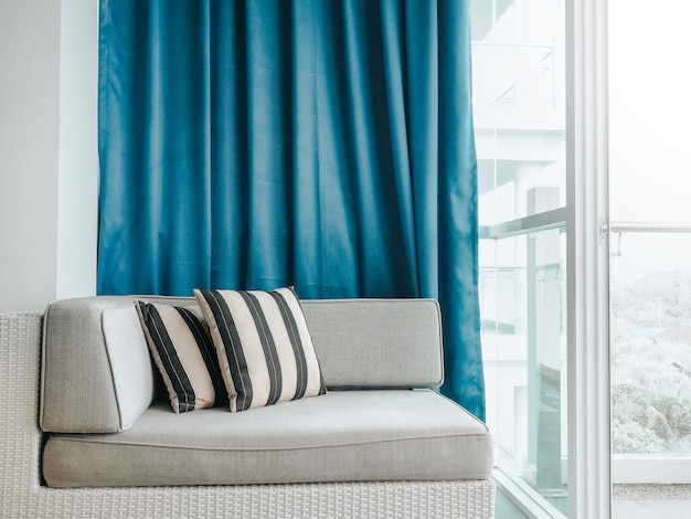 Comfortabele rotan slaapbank met kussens versierd op balkon en terras op hoog gebouw op blauw gordijn bij glazen deur in de kamer.