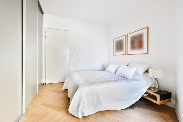 Comfortable bed with white linen and cushions in light room with frames on wall and built in wardrobe