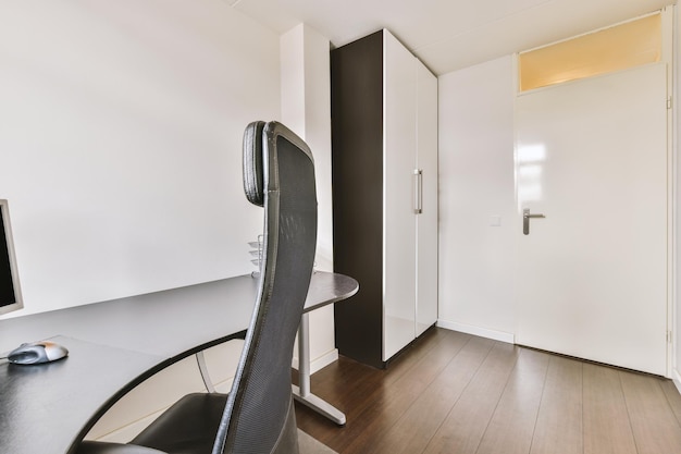 Comfortable chair placed near desk with computer monitor and\
wardrobe close to the door in light workplace in modern\
apartment