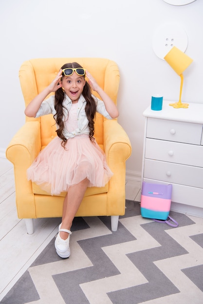 Comfortable place to rest. Girl sit chair fashionable outfit. Child girl happy face relax on shopping. Fashionista excited about shopping. Kids boutique fashion clothes salon. She is fashion addicted.