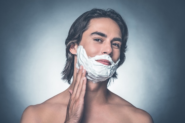 Comfortable shaving. Handsome young shirtless man spreading shaving cream over face and looking at camera while standing against grey background