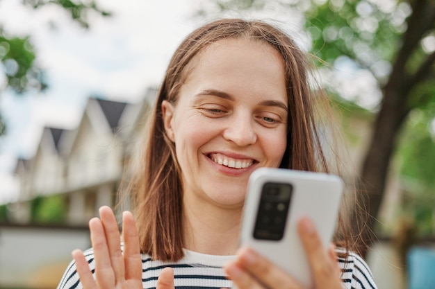 Foto communicatie op afstand jonge volwassen vrouw met smartphone die buiten video-oproep maakt glimlachende vrouw zwaaiende hand naar mobiele telefooncamera terwijl ze op straat loopt