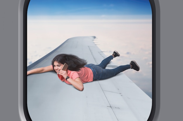 Communicatie verslaving, jonge vrouw met telefoon ligt op vliegtuigvleugel tijdens de vlucht, lucht en wolken op de achtergrond