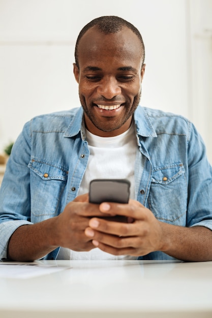 Communicating. Attractive alert dark-haired afro-american man smiling and writing a message on his phone while sitting at the table