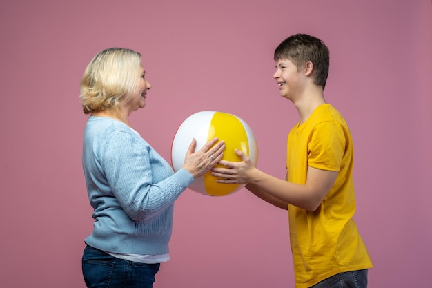 Communication. Joyful guy with down syndrome and middle aged woman standing sideways to camera opposite each other holding ball together
