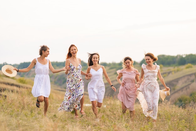 The company of cheerful female friends have a great time together on a picnic in a picturesque place overlooking the green hills. Girls in white dresses dancing in the field