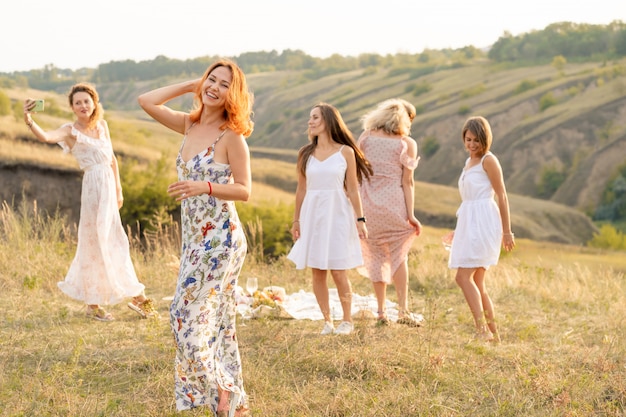 The company of happy female friends having fun and dancing outside in a picnic at hills.