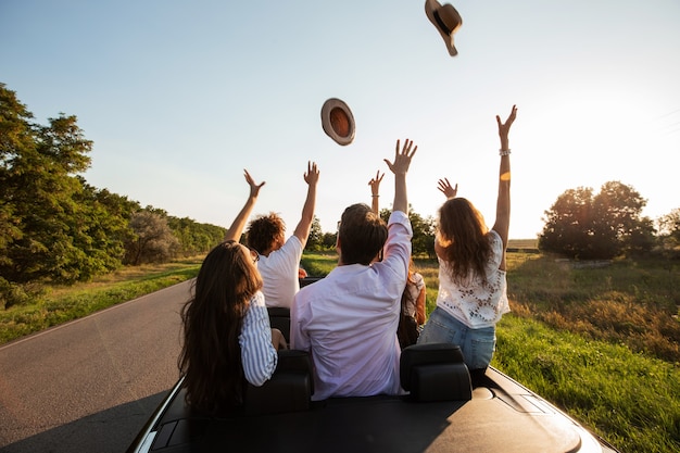 Company of happy young girls and guys sit in a black cabriolet road and throw up their hats on a sunny day. .