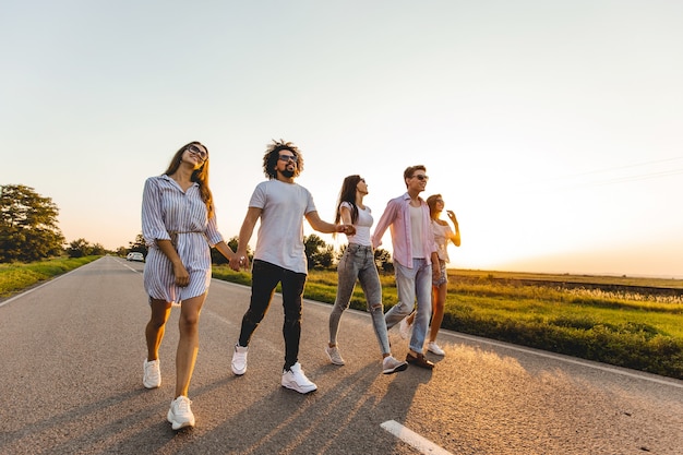 Photo company of happy young stylish guys hold their hands and walk on a country road on a sunny day .