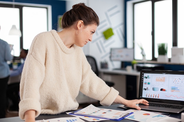 Company manager reading, analysing statistics on laptop in start up office. Executive entrepreneur, manager leader standing working on projects with diverse colleagues. Successful corporate profession