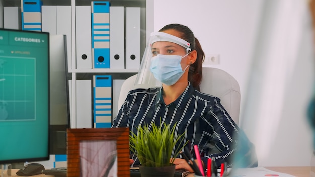 Company manager with face mask typing on computer and discussing with coworker during coronavirus pandemic in new normal business office respecting social distance. Freelancer using modern technology