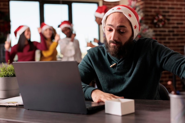 Работник компании чувствует себя обеспокоенным на работе в офисе из-за шумных коллег, празднующих канун Рождества. Усталый раздраженный сотрудник перегружен и работает во время зимнего курортного сезона.
