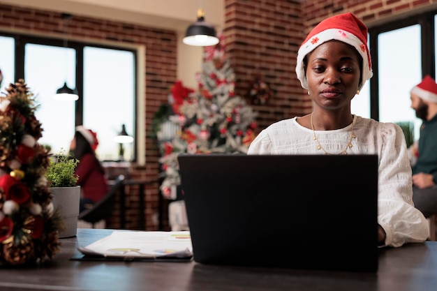 Company worker using laptop at startup office, celebrating winter season with christmas decorations and tree. Woman with santa hat working in space with festive seasonal ornaments.