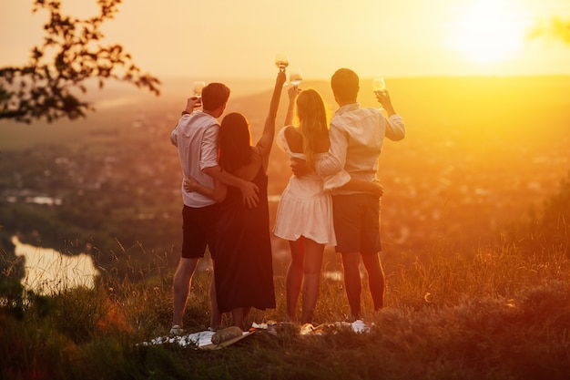 company of young friends celebrating at picnic at sunset. they standing with glasses of white wine ad enjoying perfect sunset and landscape.