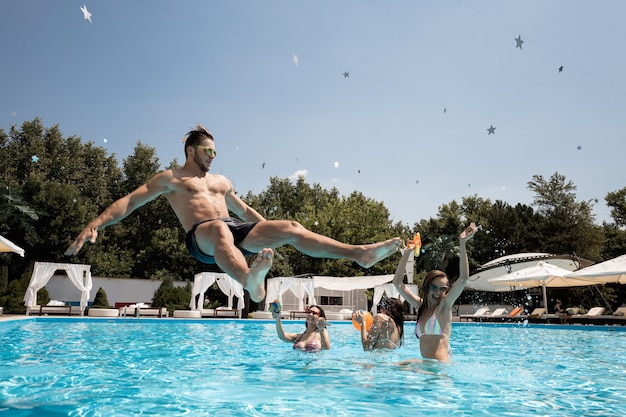 Company of young merry girls and guy relaxing in the swimming pool jumping in the water and playing with water guns on the open air on a sunny summer day next to lounge zone