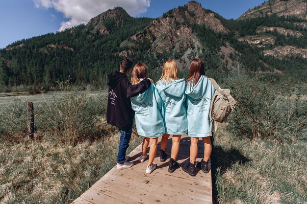 A company of young people are engaged in active recreation. The girls and the guy stand on a wooden board with their backs and look at the amazing mountain nature. A friendly hike.