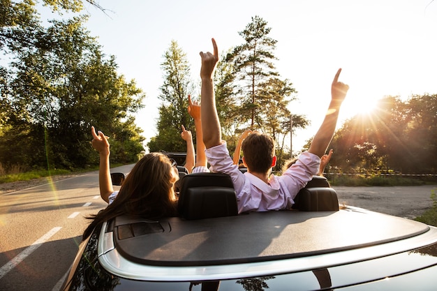 Company of young people riding in a cabriolet on the road and holding their hands up on a warm sunny day. Back view. .