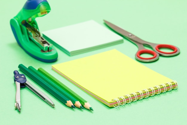 Photo compass, color pencils, notebook, note paper, stapler and scissors on green background.