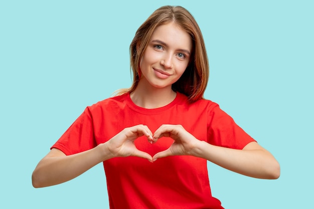 思いやりのサイン愛のケア青い背景に分離されたハートのジェスチャーを示す赤いTシャツの支持的な陽気な女性の肖像励ましの賞賛ロマンチックなメッセージ