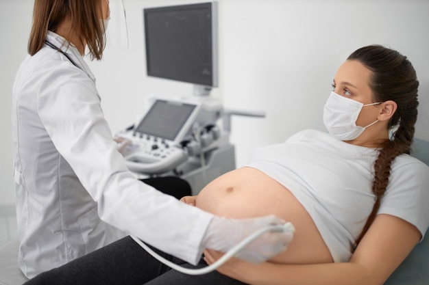 Competent female doctor in medical protective mask doing ultrasound screening for pregnant woman at modern clinic