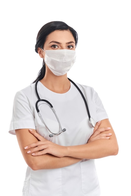 Competent female doctor wearing medical uniform and face mask standing with crossed arms on white