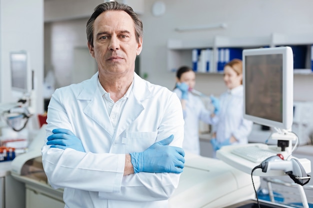 Competent man. Gray-haired practitioner pressing lips and looking straight at camera while standing isolated on lab background