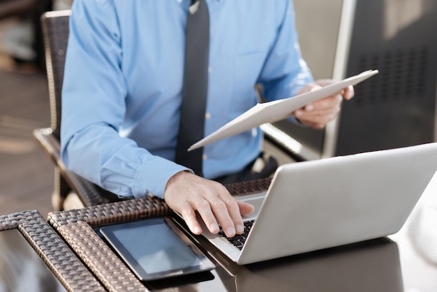 Competent manager holding folder with documents in left hand and typing text while using keyboard of his laptop