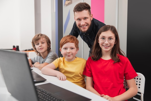 Giovane insegnante maschio competente e bambini felici che guardano la telecamera durante la lezione in aula moderna con laptop a scuola