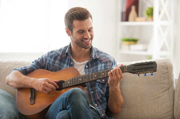 Componeren van zijn nieuwe nummer. Gelukkige jonge man die gitaar speelt terwijl hij thuis op de bank zit