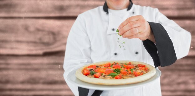 Composite image of portrait of a chef holding a pizza and adding herbs