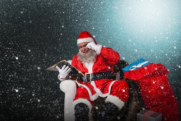 Composite image of santa reading bible with sack of christmas present beside him