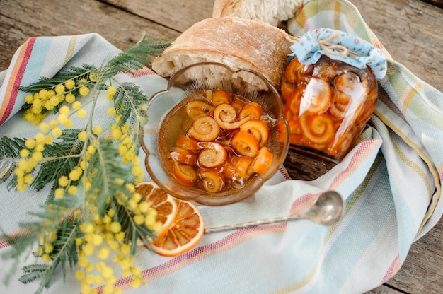 Photo composition of the candied orange spiral peel with sugar syrup in a glass jar and plate near the saucer with walnuts, mimosa, spoon and bread on the napkin on the wooden table