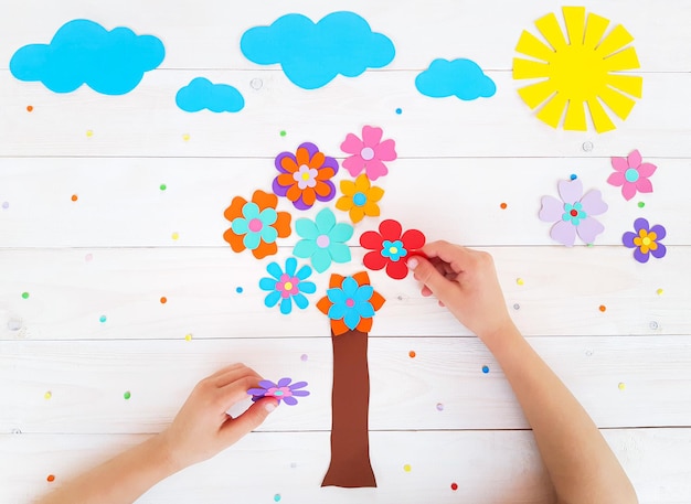 Composition of colorful flowers, sun, clouds from multi-colored paper, step by step. Tree of flowers. Child make crafts his own hands. DIY handmade art creativity on wooden table. Top view, copy space