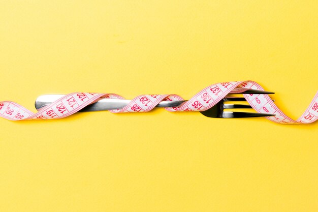 Composition of curled wrapped measuring tape and fork on yellow background.