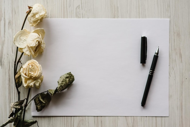 Composition dried rose flowers sheet of paper and pen for message on a white wooden background Postcard with copy space Frame Place for text design Greeting card Flat lay top view