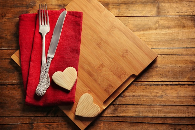 Composition of fork knife napkin and decorative hearts on cutting board on wooden table background