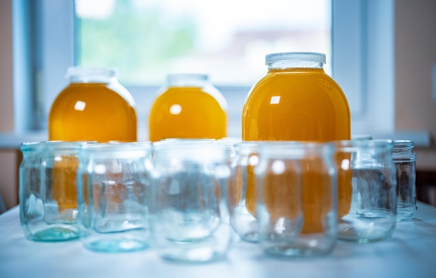 Composition of a large number of transparent empty glass jars and three large glass transparent jars with yellow sweet honey, standing on a large white table against a background of bright sunlight