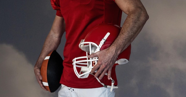 Composition of male american football player holding ball and helmet over blue sky with clouds