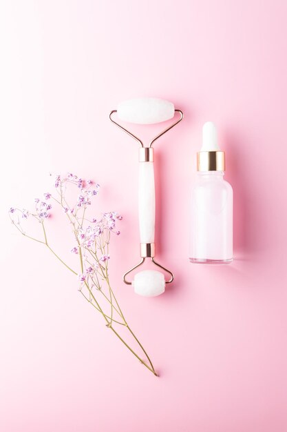 Composition of mockup serum bottle, pink quartz stone face roller with gipsophyla flowers on pink background. Natural cosmetics concept. Creative cosmetics flat lay.