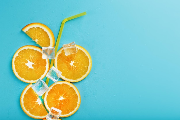 A composition of orange slices and ice cubes with a straw on a blue background in the form of a refreshing drink. Top view, free space