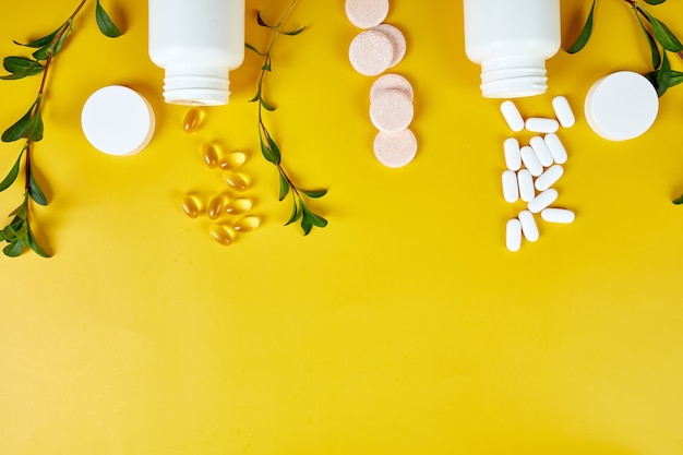 Composition of pills, fish oil, vitamins with green leavws on yellow surface