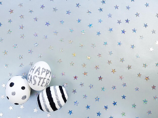 Photo composition of silver glitter inscription happy easter on a white egg and metallic stars spangles sequins. eggs with black and silver lines and polka dots. flat lay, top view, close up, copy space