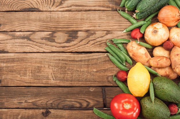 Composition with assorted fresh organic vegetables and fruits. Cucumbers, tomatoes, radish, avocado, peas, potatoes, lemon, onions. Food on dark wooden surface.