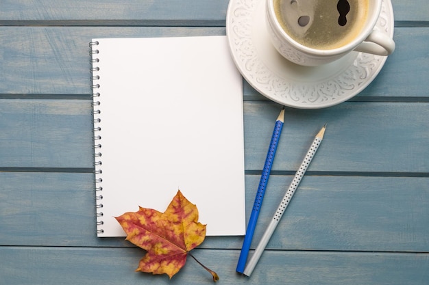 秋の構成は、青い木製のテーブルの上に空白のノートブック一杯のコーヒーを残します