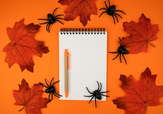 Composition with blank notepad pen autumn leaves and spiders on orange background Halloween