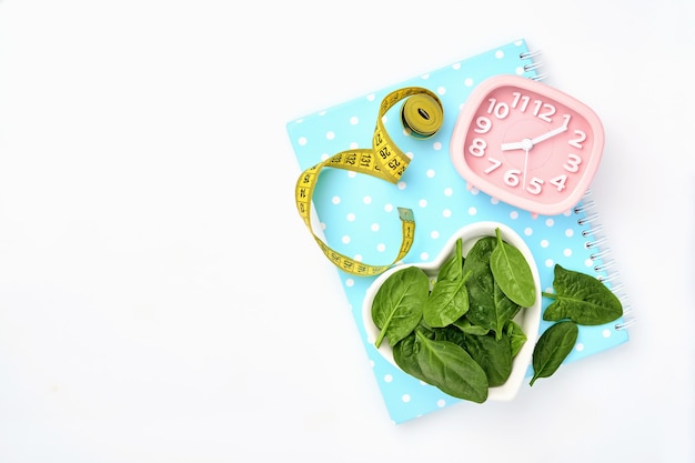 Composition with  spinach, measuring tape, blank paper notebook and pink alarm clock on white table.  Diet concept, copy space